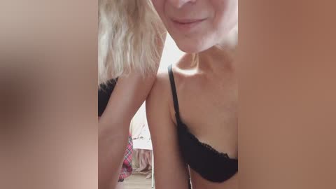 A video of a blonde woman with fair skin and a slender build, smiling, wearing a black lace bra. A partially visible person in a red plaid shirt is behind her.