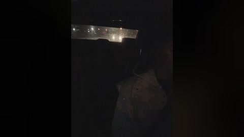 A dimly lit, grainy video of a darkened room with a man in silhouette, holding a flashlight, revealing a distant, bright scene of a cityscape with numerous illuminated buildings and lights.