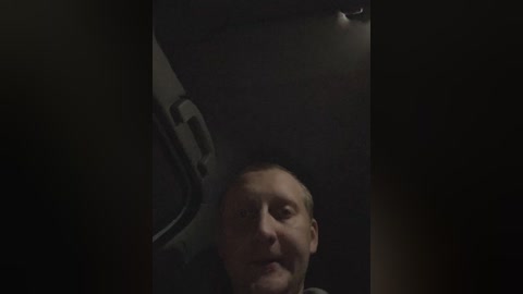A dimly lit video captures a bald, middle-aged man in a car, looking upwards. His face is partially obscured by the car's interior, with a dark background and a faint glimpse of a vehicle door handle.