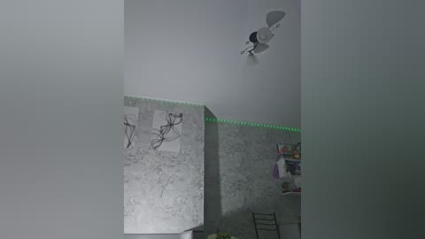 A video of a dimly lit, minimalist bedroom with a white ceiling fan, a green LED strip, and two black-and-white sketches on a marble-patterned wall. A chair and a small table with a purple cloth are visible in the foreground.
