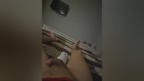 Video of a man with a large, erect penis, lying on a bed with a beige and brown striped blanket. He is holding a white vibrator and a red sock, in a dimly lit room with a flat-screen TV mounted on the wall.