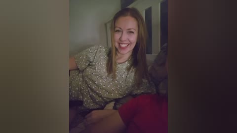 Video of a smiling, light-skinned woman with straight, shoulder-length light brown hair, wearing a floral-patterned shirt, sitting on a bed in a dimly lit room.