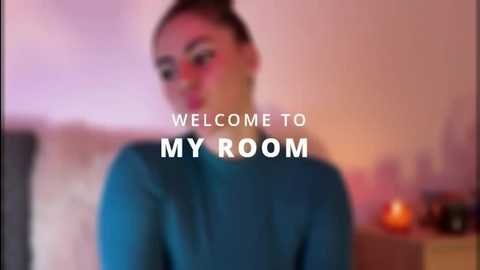 Video of a woman with light skin and brown hair in a ponytail, wearing a teal sweater, with blurred background text reading \"Welcome to My Room.\