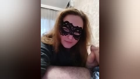 Video of a fair-skinned woman with long blonde hair, wearing a black lace masquerade mask, lying on a bed, with a man's hairy arm in the foreground.