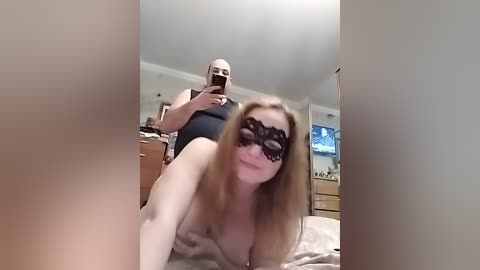 Video of a fair-skinned woman with long blonde hair wearing a black lace mask, lying on a bed in a bedroom. A bald, muscular man is behind her, holding a phone.