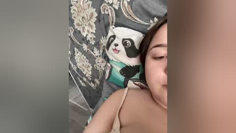 A video of an Asian woman with light skin and straight, brown hair partially covering her face. She wears a beige spaghetti strap top and rests her head on a pillow with a panda design.