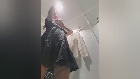 A video of a woman in a bathroom, wearing a black dress, lifting her arms. She holds a red object in her hand. A white towel hangs behind her on a hook.