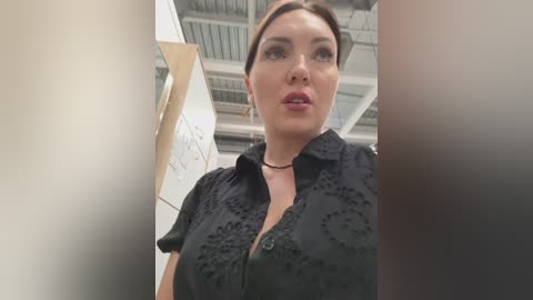 Video of a middle-aged woman with pale skin and brown hair, wearing a black floral-patterned blouse, standing in a white-walled room with exposed ceiling beams and a wooden board.