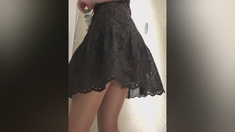 Video of a woman wearing a black lace dress, revealing her bare legs and thighs. The background shows a beige curtain with a subtle geometric pattern.
