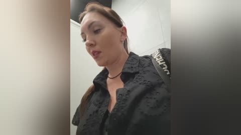 Video of a fair-skinned, brunette woman in a black lace top, sitting in a tiled bathroom, wearing a black choker. She has a neutral expression.