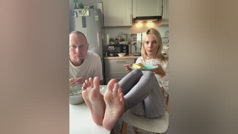 Video of a blonde woman in a white top and grey leggings, sitting on a white stool in a modern kitchen, holding a plate, while a bald man in a white shirt sits next to her.
