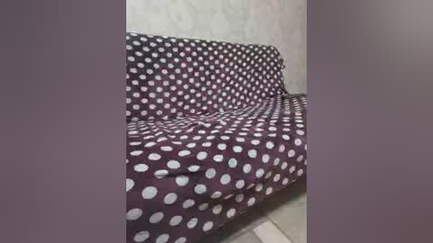 A video shows a small, dark purple sofa with a white polka dot pattern against a plain, light-colored wall. The sofa is positioned against a corner, with a blurred background, suggesting a simple, sparsely furnished room.