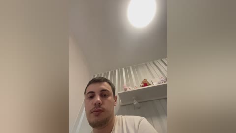 Video of a young man with short dark hair and a light beige shirt, taken from a low angle. Background includes a white curtain with a child's toy on a shelf.
