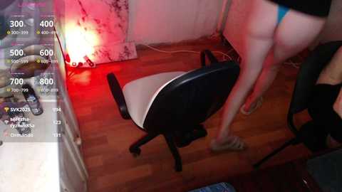 Video of a woman in a black crop top and blue thong, standing on a wooden floor next to a black chair, in a dimly lit room with a red light.