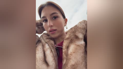 Video of a young woman with light brown hair, fair skin, and brown eyes, wearing a luxurious brown fur coat, posing against a blurred background.