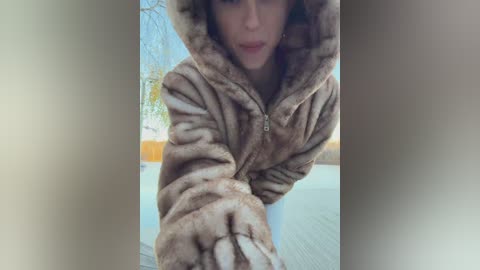 Video of a woman wearing a brown fur-lined hooded jacket, reaching out with a gloved hand, with a blurry background suggesting an outdoor setting.