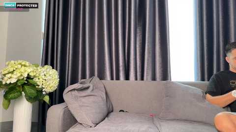 Video of a man in a black t-shirt sitting on a grey sofa, with dark grey drapes in the background. A vase of white hydrangeas is on the left.