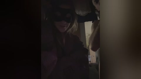 A dimly lit video shows a woman with blonde hair and a black mask, peering through a window. The background is indistinct, casting a mysterious, suspenseful atmosphere.