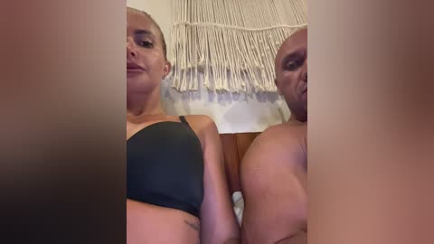 Video of a dark-skinned man and light-skinned woman in a bedroom, both shirtless, with a beige macram\u00e9 wall hanging in the background.