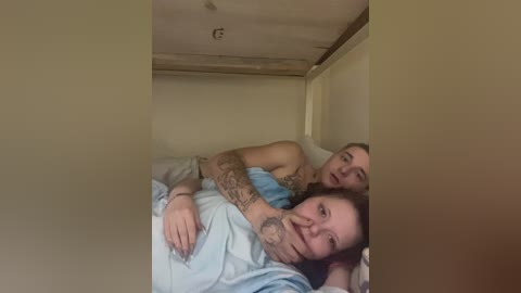 A video shows a young couple lying in a small, dimly lit room with beige walls, both wearing light blue hospital gowns. The woman has tattoos and a smile, while the man, partially obscured, appears to be looking at her.