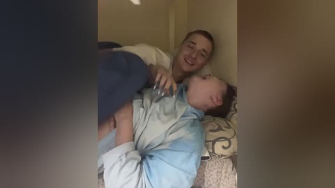 Video of a smiling young man cradling a sleeping toddler in his arms, both in pajamas, on a patterned couch in a dimly lit room.