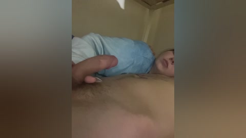 Video of a naked man with a circumcised penis and pubic hair, lying on his back, partially covered by a light blue shirt, in a dimly lit room.