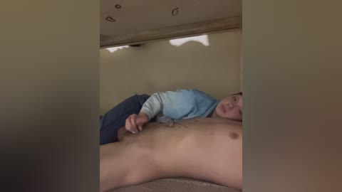 Video of a young, fair-skinned man with short brown hair lying nude on a bed, wearing only a blue hoodie and blue jeans, with a neutral expression. The room's beige walls and dim lighting suggest a private, intimate setting.