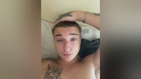 Video of a young, shirtless man with short hair and tattoos, lying on a bed, resting his head on a pillow, appearing relaxed.