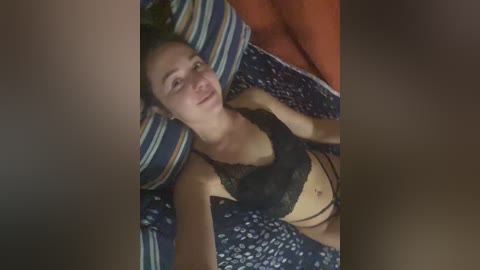 Video of a young woman with light skin, small breasts, and dark hair lying on a bed with striped pillows, wearing black lace lingerie.