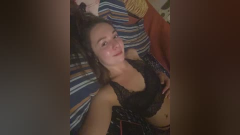 Video of a smiling, fair-skinned woman with curly hair, wearing a black lace bra, lying on a striped bed.