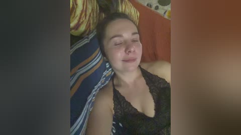 Video of a young woman with fair skin, closed eyes, lying on a bed with striped and patterned pillows. She wears a black lace camisole. Background includes orange and floral-patterned curtains.