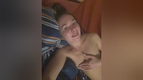 A video of a young, topless woman with light skin, brown hair, and a slender physique, lying on a bed with blue and orange pillows, smiling.