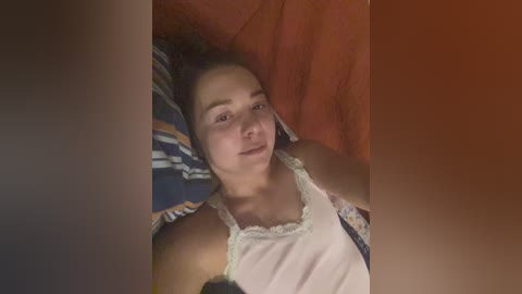 Video of a young woman with curly dark hair, lying on a bed with a red pillow and blue-striped sheets, wearing a white tank top, looking relaxed.