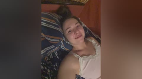 Video of a young Caucasian woman with light skin, lying on a bed with striped pillows, wearing a white tank top and smiling.