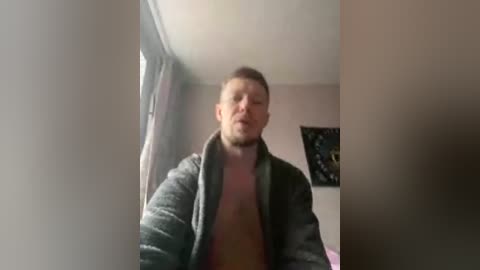 A video of a man with a beard, wearing a dark jacket, standing in a dimly lit room with beige walls, a window with curtains, and a black poster in the background.