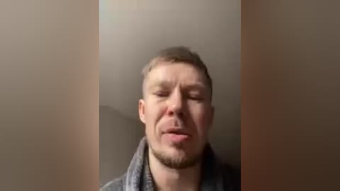 Video of a man with short, light brown hair, wearing a gray shirt, smiling with closed eyes, standing between two beige walls.