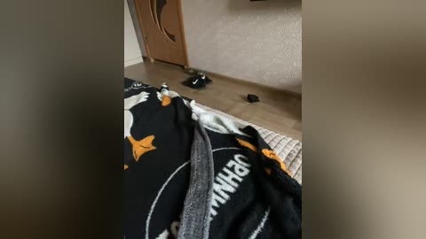Video of a messy bedroom with a black comforter featuring an orange star and planet design, a pair of black socks, and a wooden door.