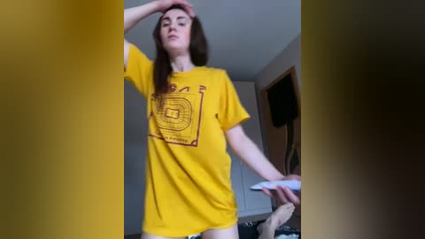 Video of a young, fair-skinned woman with long brown hair wearing a yellow T-shirt and beige pants. She stands in a dimly lit room with a flat-screen TV on the wall and a door partially open.