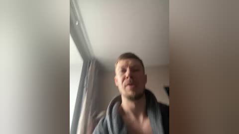 A blurry video of a young, light-skinned man with short, light brown hair, wearing a dark hoodie, standing near a window in a dimly lit room.