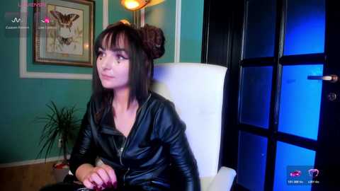 A video of a young woman with dark hair styled in two buns, wearing a black leather jacket, sitting in a white chair, in a teal-painted room with a framed picture on the wall.