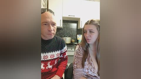 Video of a middle-aged man in a red sweater, wearing sunglasses, and a woman in a pink sweater sitting in a modern kitchen with white cabinets and a black countertop.