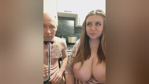A video shows a bald man and a woman with long brown hair, both topless, standing in a kitchen. The man wears a pink shirt, and the woman has large breasts.
