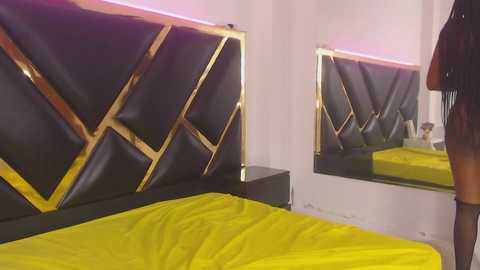 Video of a modern bedroom with a black leather bed featuring diamond-shaped gold accents, bright yellow bedding, and a mirrored wall.