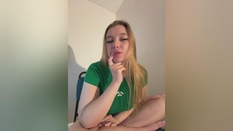 Video of a fair-skinned, blonde-haired young woman with medium-sized breasts, wearing a green t-shirt, sitting on a blue chair, leaning against a beige wall, licking her index finger suggestively.