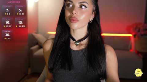 A video of a young woman with long black hair, wearing a black top and necklace, in a modern living room with a white couch and red lighting.