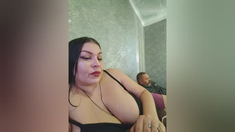 Video of a plus-sized woman with large breasts, wearing a black dress, seated in a dimly lit room with ornate wallpaper. A man with a beard and glasses sits nearby.