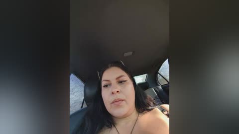 A video of a young woman with long black hair and a neutral expression, sitting in the back seat of a car.