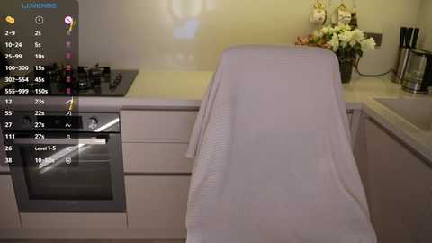 A video of a modern kitchen with a white tablecloth-covered chair in the foreground, a gas stove with a black top on the left, and a white countertop with a flower arrangement on the right.