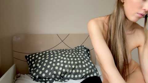 Video of a young, nude, fair-skinned woman with long, straight brown hair, seated on a bed with a beige headboard and polka-dotted pillows.