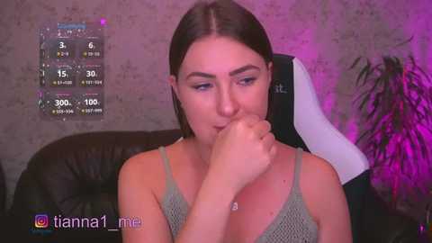 Video of a young woman with long brown hair, wearing a green spaghetti-strap top, biting her finger, seated on a black leather couch against a textured, beige wall with purple ambient lighting.